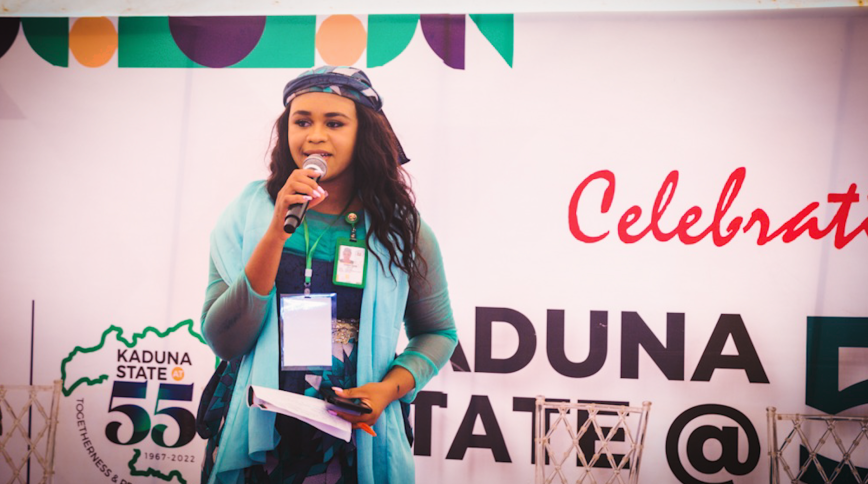 A young light-skinned Black woman with long hair speaks into a microphone on a stage