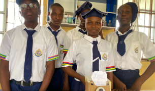 Four Nigerian teens in school uniforms stand behind a camera made out of a cardboard box and other materials